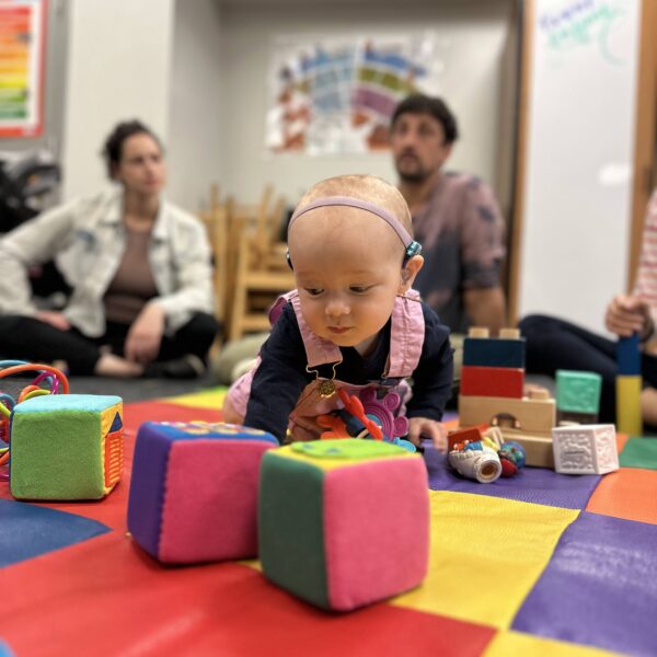 A baby with hearing aids crawls on the floor with colorful blocks. Adults sit in the background.