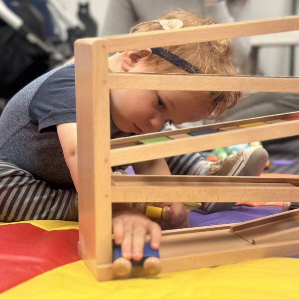 A toddler with a hearing device plays with a wooden toy while sitting on a colorful floor.